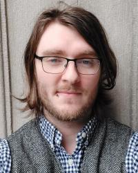 A photograph of a person with shoulder-length brown hair, a short beard, and rectangular glasses. They are wearing a checkered button-up shirt underneath a grey herringbone wool vest.