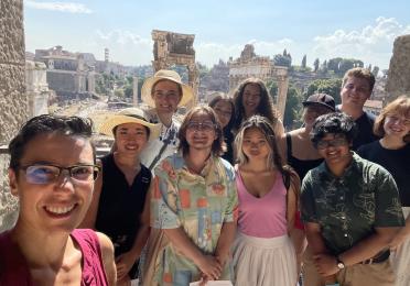 11 individuals stand in front of the ruins of the Roman Forum